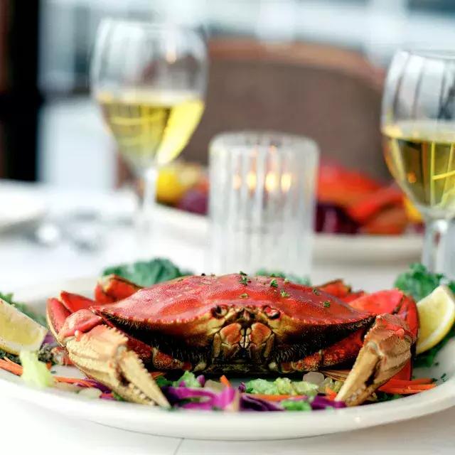 Um caranguejo Dungeness sentado em um prato em um restaurante 和 duas taças de vinho branco ao fundo.