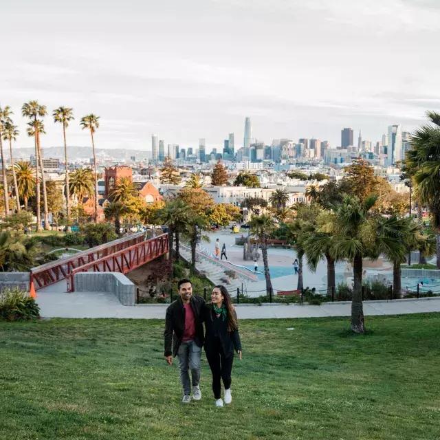 A couple walks toward the camera with Dolores Park 和 the San Francisco Skyline behind them.